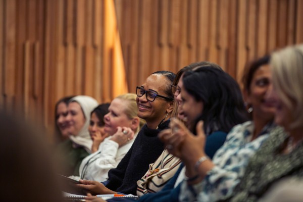 Row of women of different ages and ethnicities smiling and clapping indoors at an event.