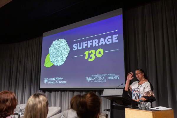 Anahera Morehu speaks at podium, suffrage 130 projected onto screen in auditorium