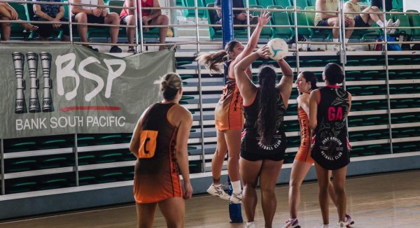 Five girls playing netball at an indoor tournament. One girl in black aims to shoot the ball with another girl in orange in front of her jumping to defend the ball. Three others stand around