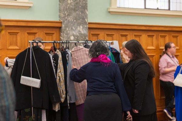 Two women look through the clothes hanging on a rack at the dress for success event. They have pulled out a pink checkered blazer and are looking at it. 