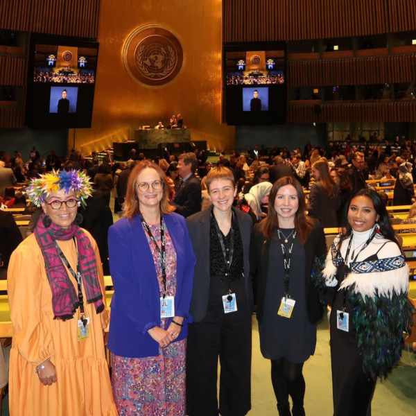 Five women stand together inside a wooden building (UN HQ, New York). Women are dressed in bright clothes and smiling. 