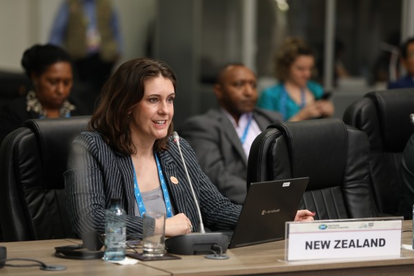 Woman sits at a desk with a New Zealand sign in front and speaks into a microphone.