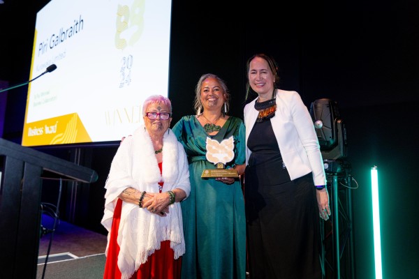 Three maaori women stand on stage smiling. The woman in the middle is in green and holding an award.