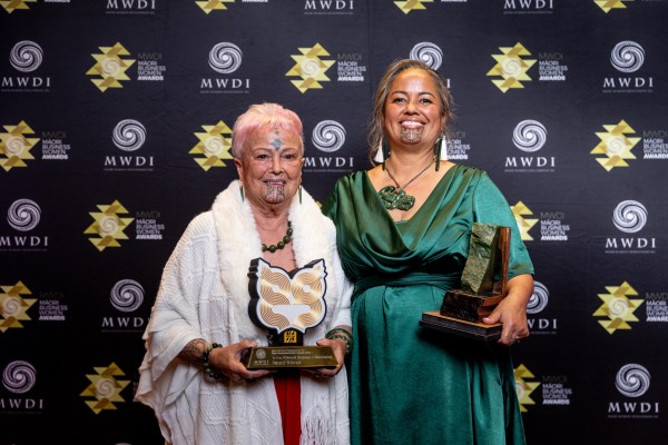 Two maori women hold awards and smile in front of a black backdrop with logos